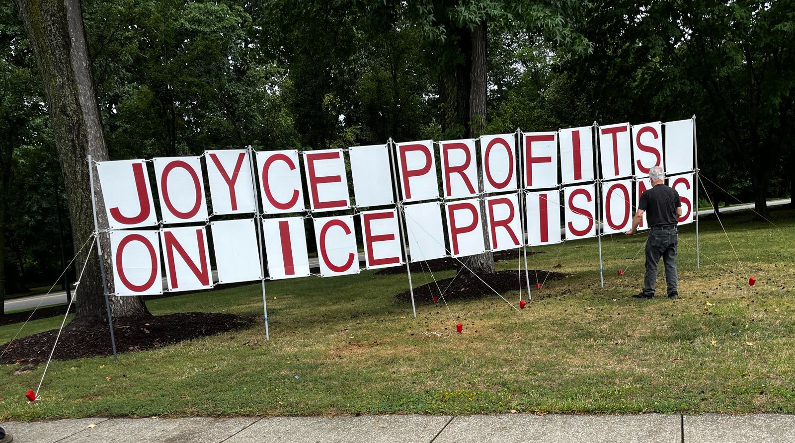 Ohio District 14 Partners volunteers setting up a large letter sign reading "Joyce profits on ICE prisons" on a roadside lawn.